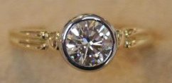 A round brilliant diamond bezel-set in a yellow-gold band, with decorative shoulder flourishes and shallow milgrain bevels either side of the bezel.